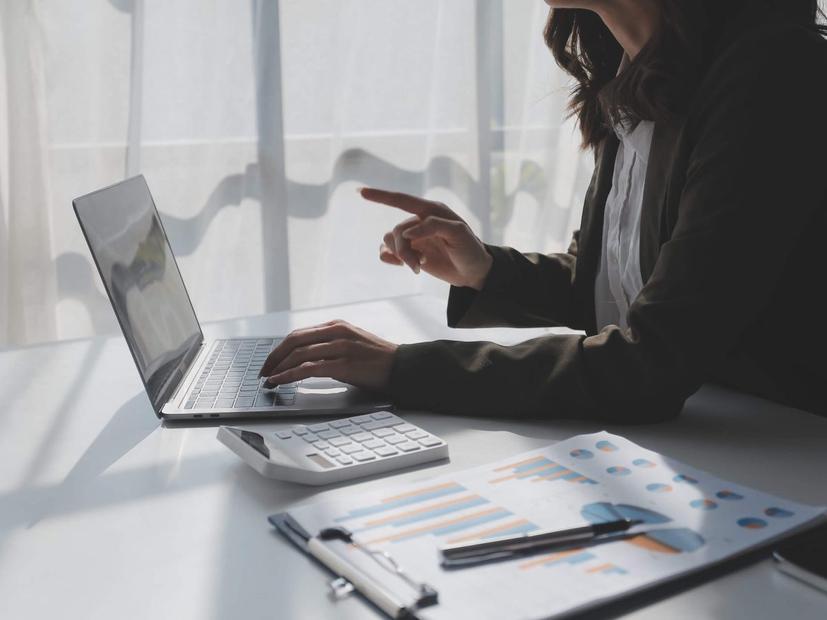 In a sunlit workspace, a professional woman engages in an important discussion, confidently navigating through her laptop while analyzing data reports spread across her desk.