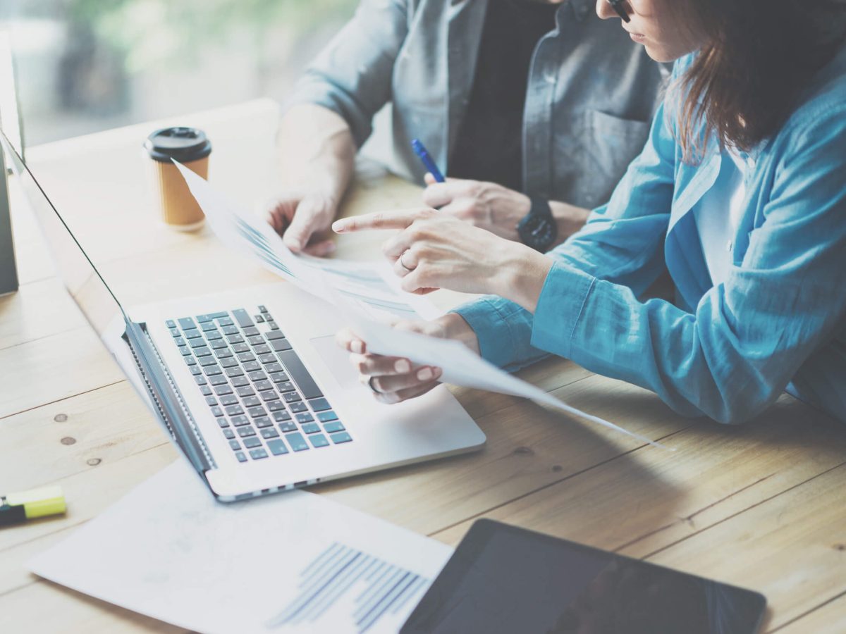 A team of professionals engages in a dynamic brainstorming session, combining ideas and insights while analyzing reports and data at a sunny workspace. The atmosphere is filled with energy as their laptops and documents lay scattered, reflecting their dedication to achieving success together.