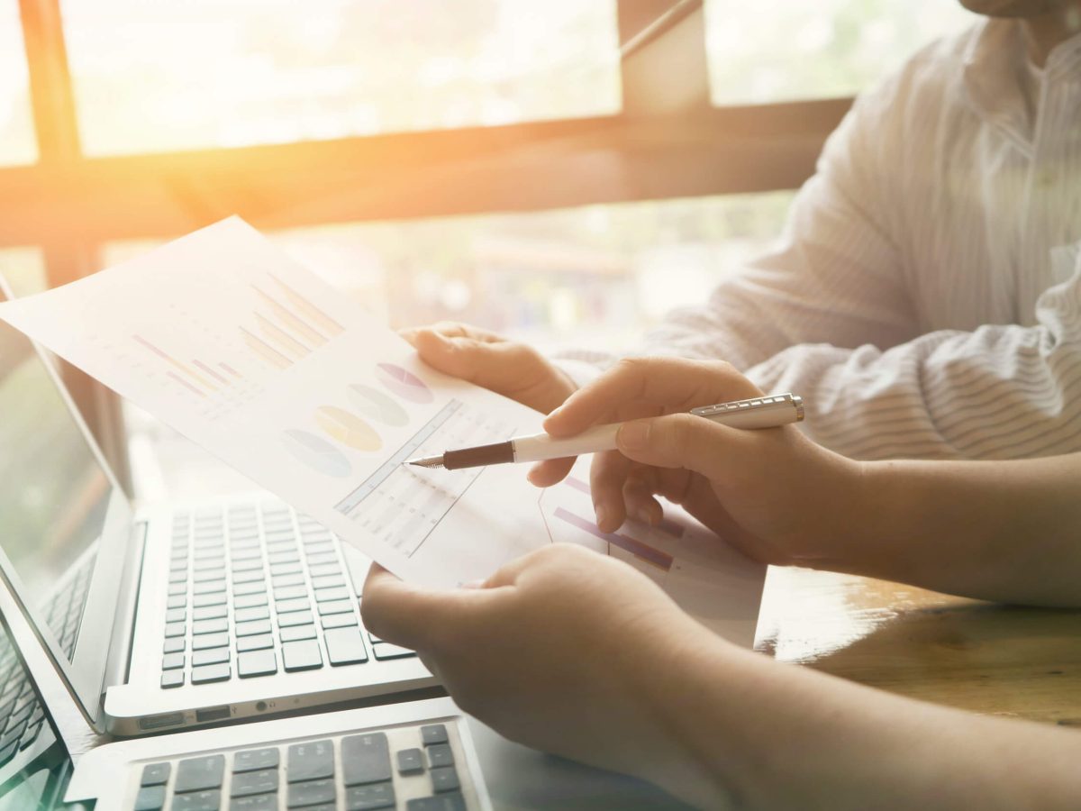 In a bright office environment, two colleagues collaborate on a project, analyzing charts and data on paper while utilizing a laptop to capture their findings. The sunlight filtering through the windows adds a warm glow, enhancing their focus and teamwork.